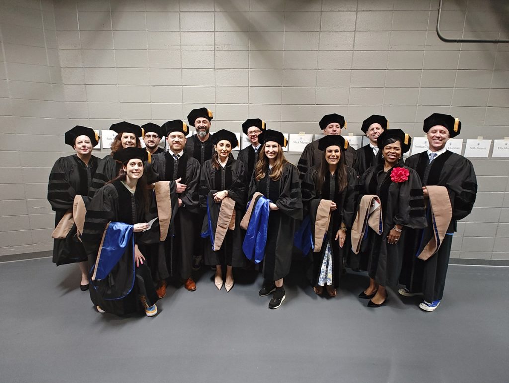 Group shot of fourteen men and women in caps and gowns who have earned the Doctor of Business Administration degree just before they walk in the 2025 Johnson & Wales Commencement ceremony in Providence.