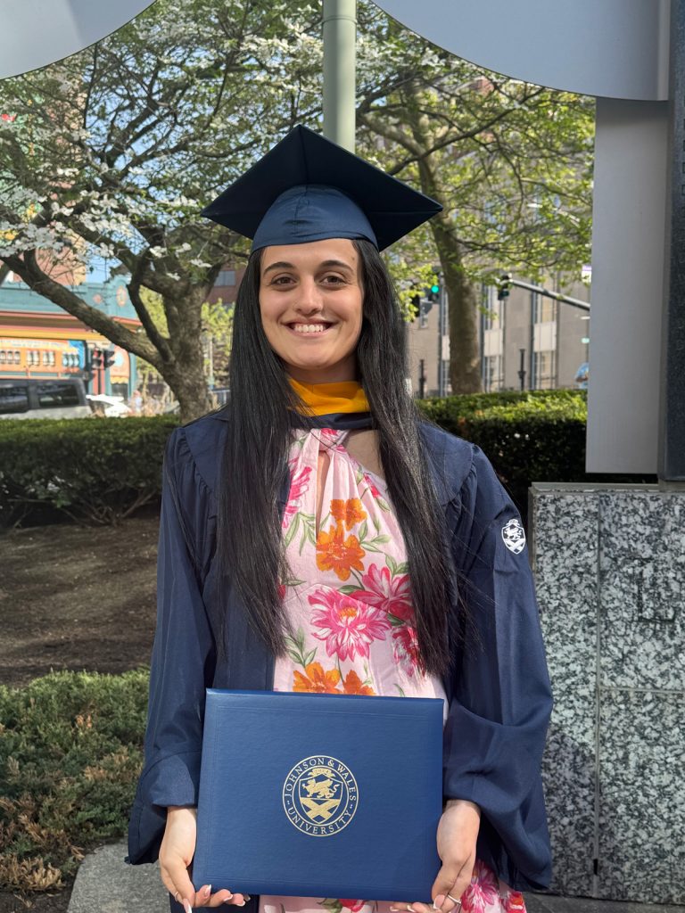 Smiling female in cap and gown, holding the diploma she received at the 2025 Johnson & Wales Commencement ceremony in Providence.