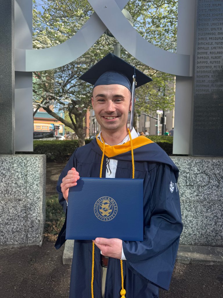 Smiling male, Ben Huntzinger, in cap and gown, holding the diploma he received in the 2025 Johnson & Wales Commencement ceremony.