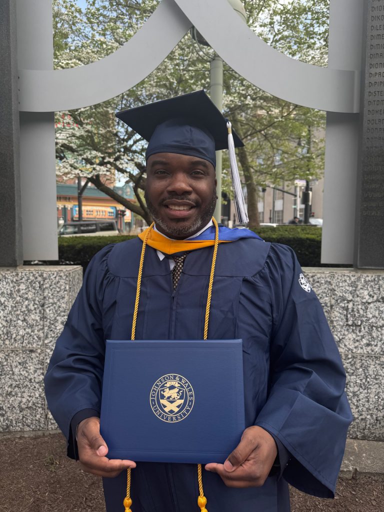 Smiling male, Chef Daniel Thomas, holding the diploma he received in the 2025 Johnson & Wales Commencement ceremony in Providence,