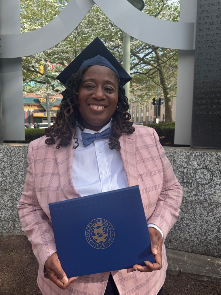 Smiling woman, Taralyn Stephens, wearing a cap, blue bowtie and pink jacket with a blue windowpane design. holding the diploma she received at the 2025 Johnson & Wales Commencement ceremony in Providence.