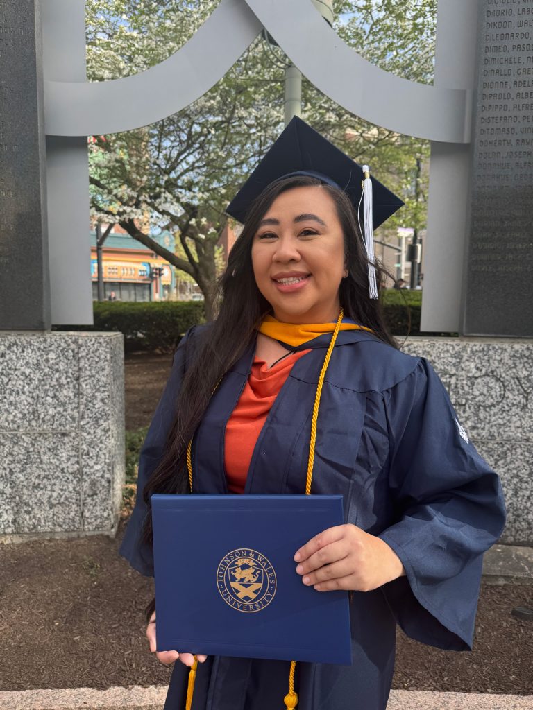 Smiling woman of Asian descent in a cap and gown, holding the diploma she received in the 2025 Johnson & Wales Commencement ceremony in Providence.