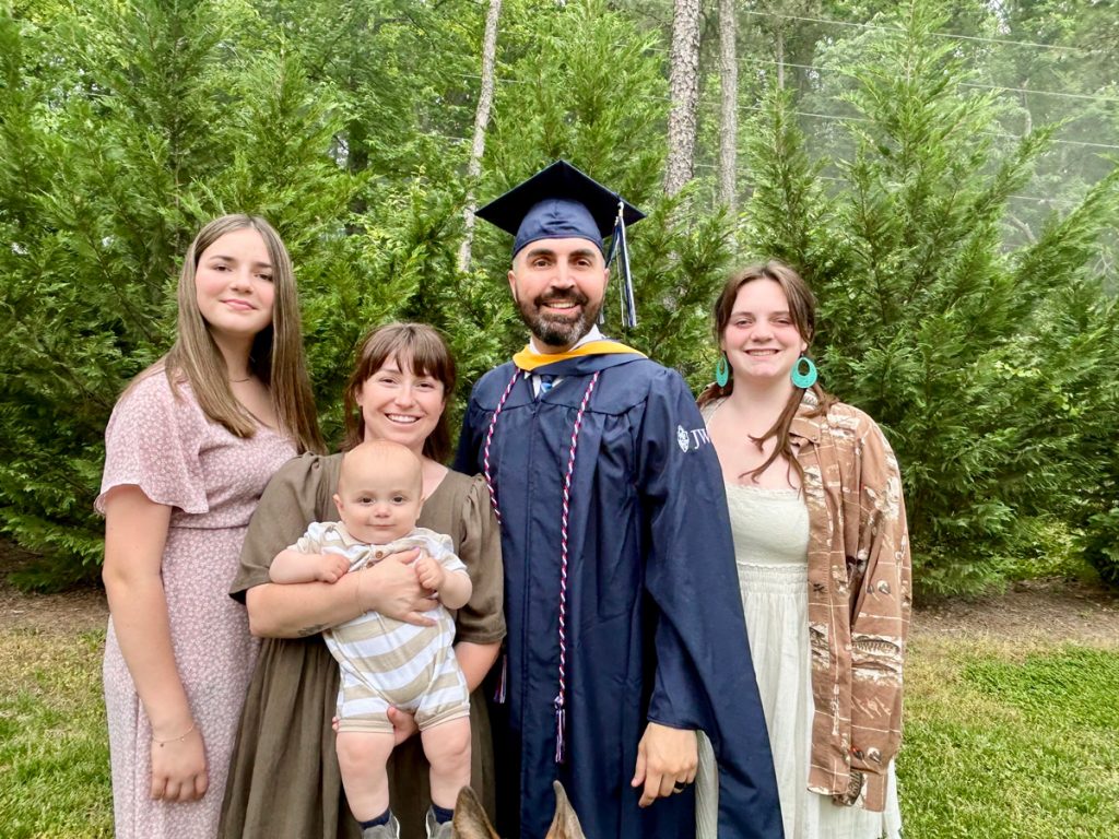 Smiling male, Jonathan Garrigues, outdoors in his cap and gown, surrounded by his two daughters and wife, who is holding their baby, on the day of the 2025 Johnson & Wales Commencement ceremony in Charlotte.