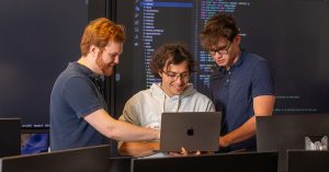 Three young men view a laptop screen together.