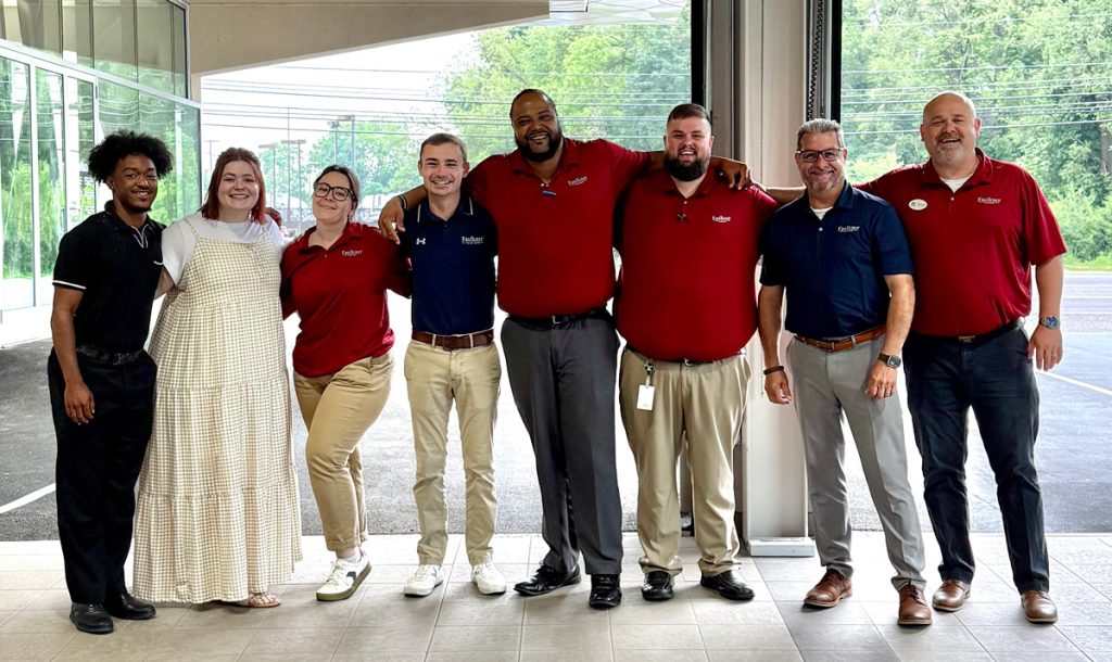 Young man, Ben, and seven co-workers of mixed gender, stand smiling and with arms around each other.