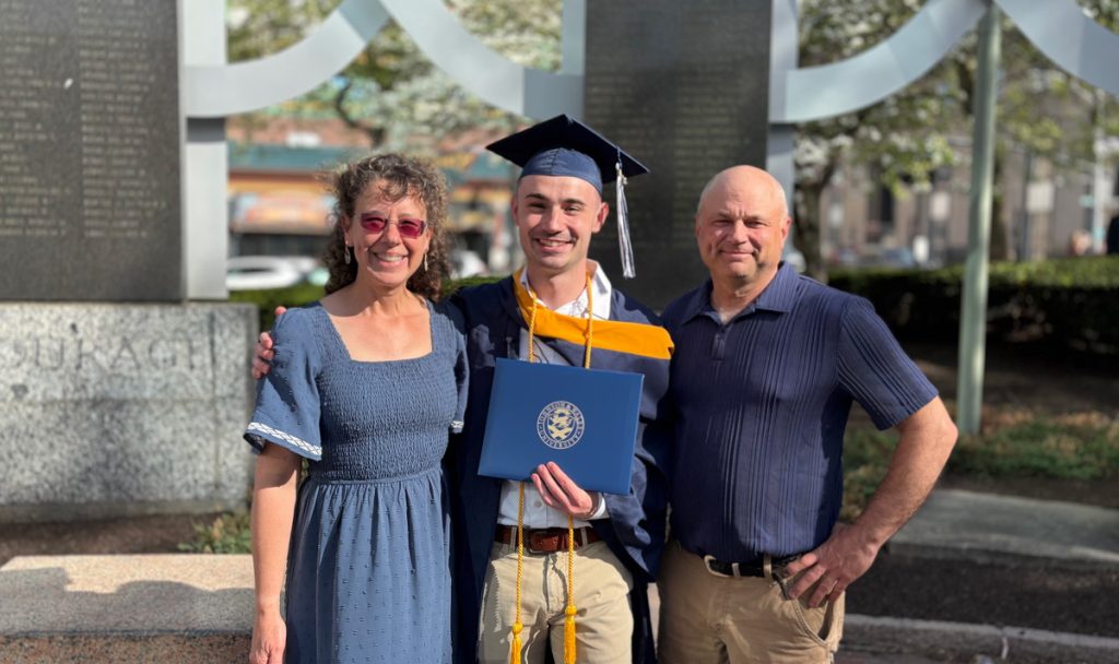 Young man, Ben, in cap and gown and holding his diploma, with his mom and dad after the JWU Commencement Ceremony in Providence.