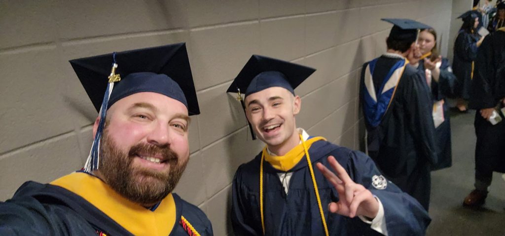 Two young men, Ben and a classmate, in caps and gowns, smiling as they wait for their commencement ceremony procession to begin.