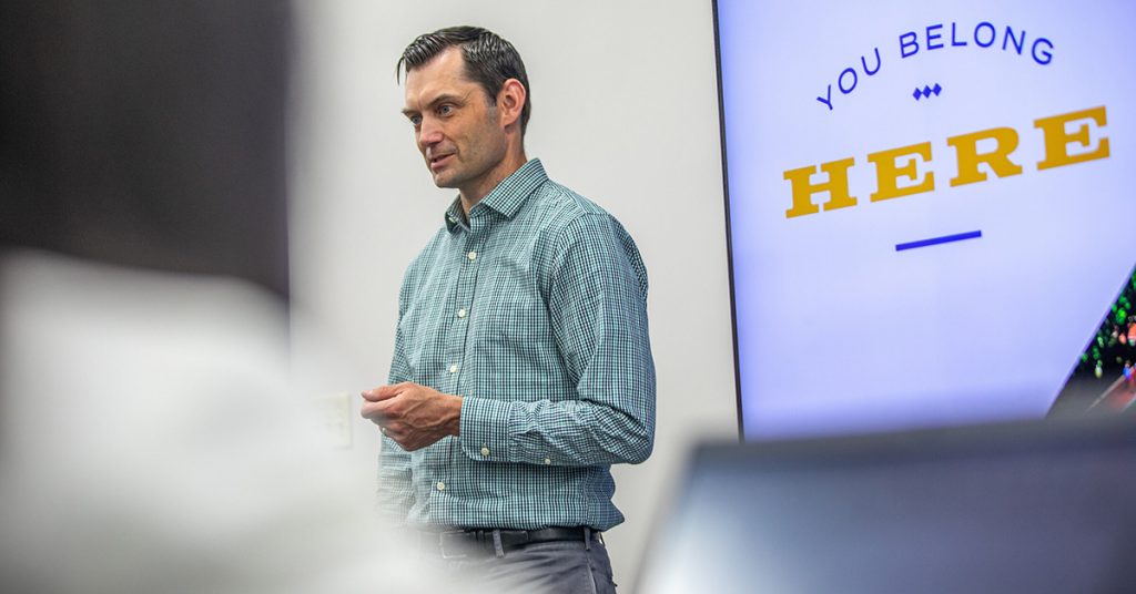 Male in a classroom making a presentation, standing in front of a screen with the words: You Belong Here.