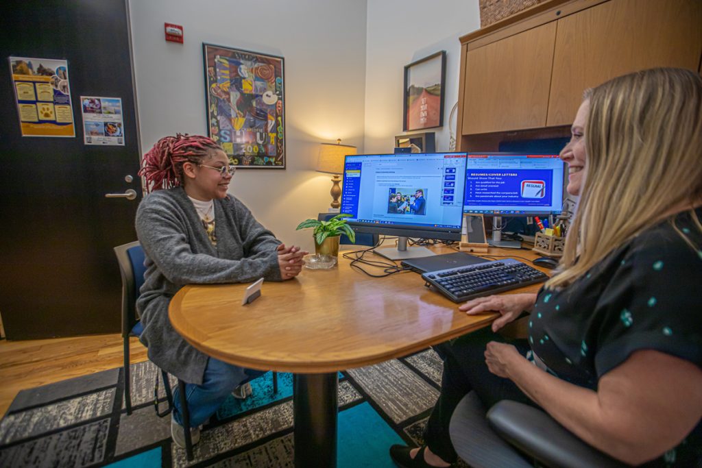 Two women seated at a table ib ab office area viewing two screens with graphic information.