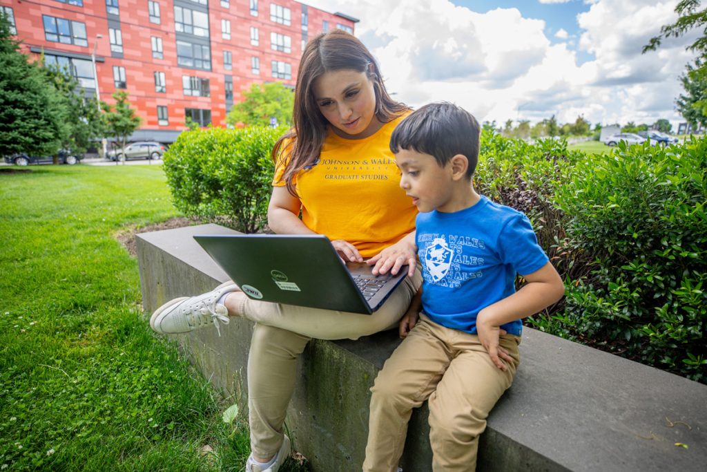 Young woman and her young son seated on a low wall in an outdoor campus area, looking together at a laptop screen.