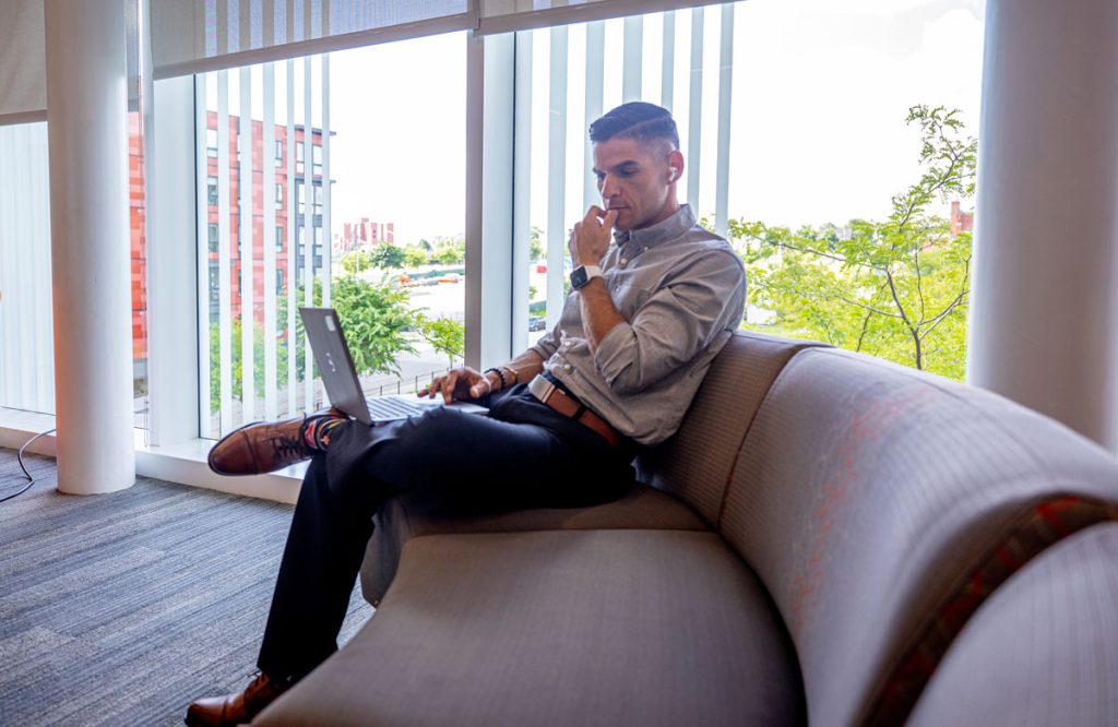 Thoughtful looking male seated on couch in a common area of a university building with a laptop on his lap, 