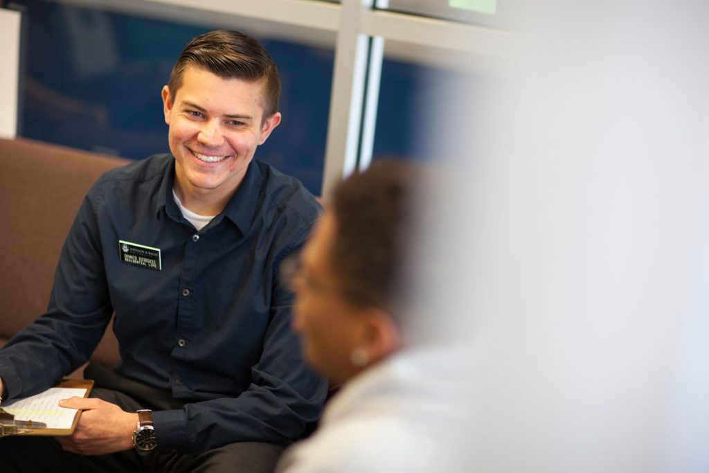 Smiling man in business casual clorting and a company name tag is seated facing a person.