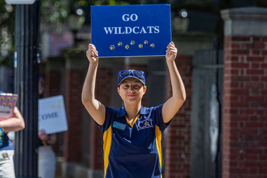 Woman wearing JWU blue and gold shirt, with sunglasses on her cap, holding a sign above her head with the words: Go Wildcat.