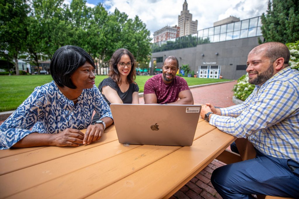 Two men and two women of mixed ethnicity seated around a table in a green space outside of university buildings, all looking at one laptop screen.