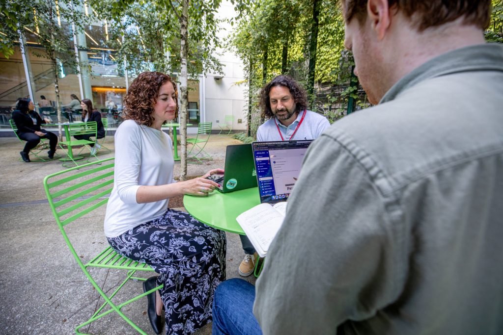 Woman and two men seated at a table in an outdoor school courtyard, viewing laptop screens.