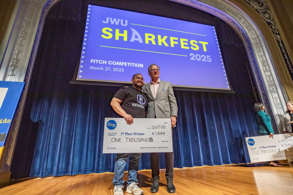 Randy Benitez. holding a cardboard "check" for $1,000 and his mentor, Dr. Jeff Binczyk, pose together onstage at the JWU Sharkfest 2025 pitch competition.