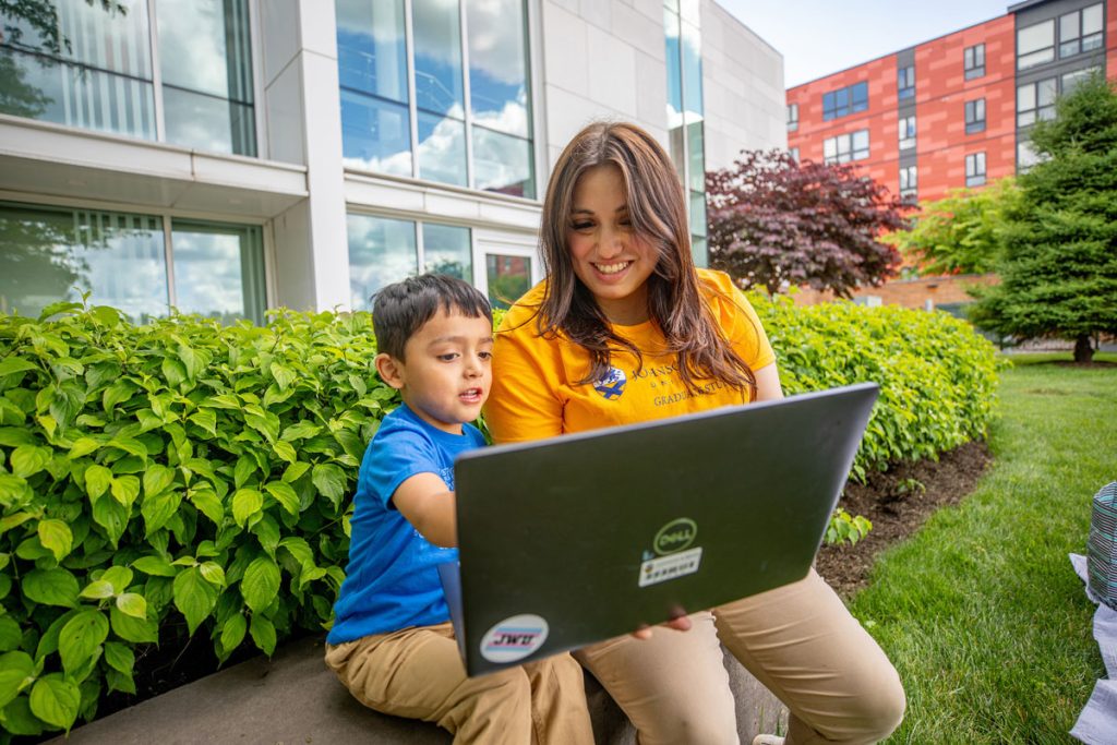 Young boy and his mom, sit outdoors in a landscaped campus area and view a laptop screen together.
