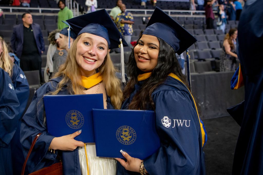 Two young women in JWU caps and gowns are holding their diplomas and smiling.