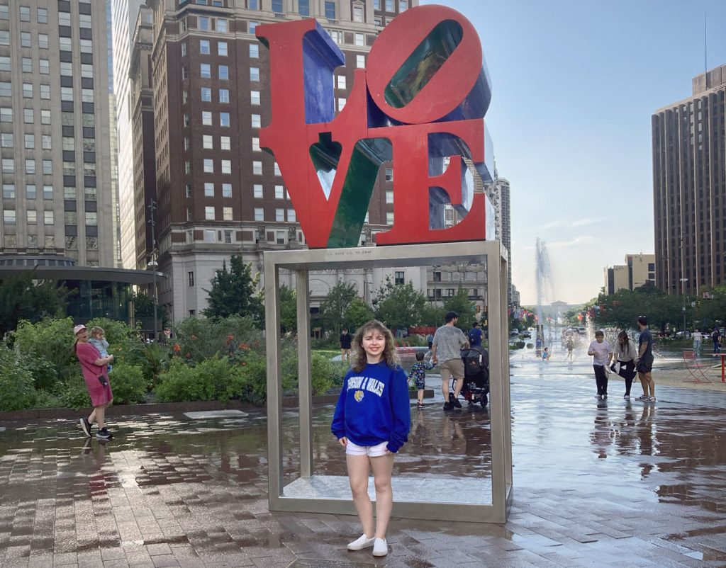 Katherine Campbell wearing her blue Johnson & Wales sweatshirt, standing in fron the LOVE sculpture in the JFK Plaza.