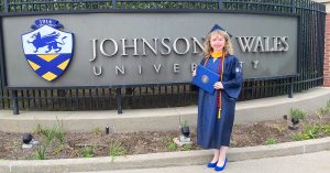 Katherine Campbell in her JWU cap and gown stands in from of the campus wall with "Johnson & Wales University" and the school logo.