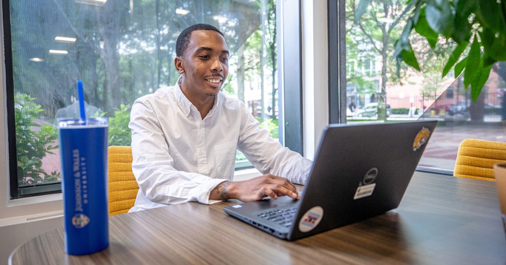 Male seated at table smiles as he views his laptop screen.