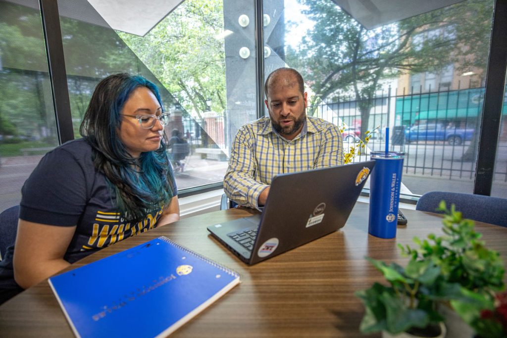 Female student and male advisor seated at a table in front of windows with a view to the street, look together at a laptop screen.