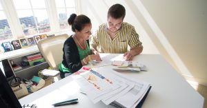 Two people at a bright office desk reviewing printed charts and papers near a computer and phone.
