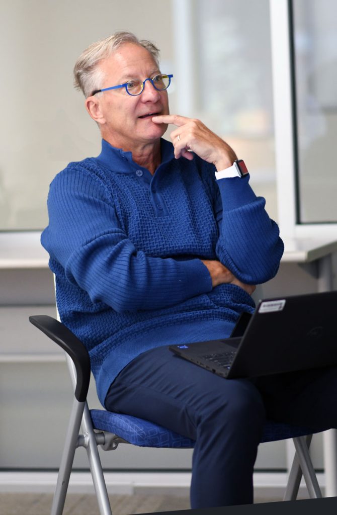 Dr. Jeff Binczyk wearing a blue sweater and seated in from of a window, looks thoughtful while listening to a student during a virtual mentoring meeting.