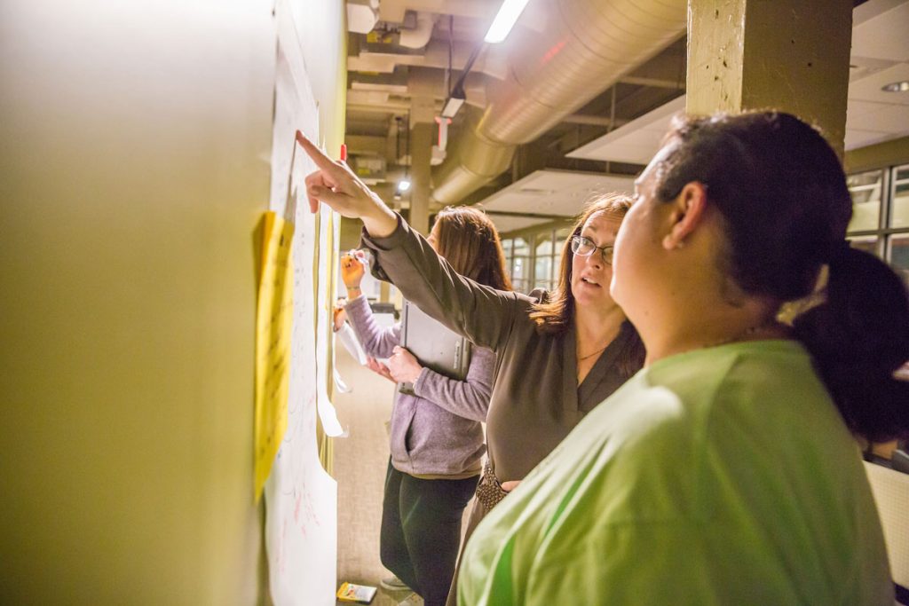 Several women, one of them pointing at a poster on the wall, inside a university building,