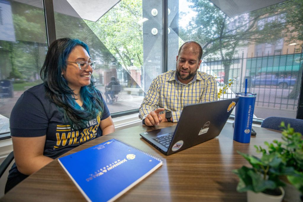 Young woman and young man seated at a table in front of windows with a view to a campus courtyard, viewing a lab top screen together.