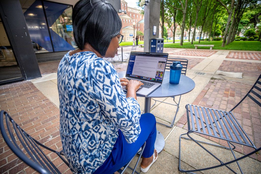 View of a laptop screen over the shoulder of a woman seated at small round table in an outdoor courtyard adjacent to a university building.