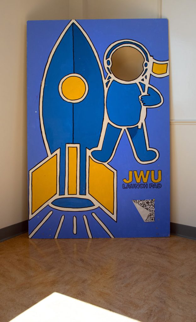 The Launch Pad poster showing an astronaut with a flag, next to a blue and yellow-gold rocket, on a blue backgrounsd with the gold JWU insignia.