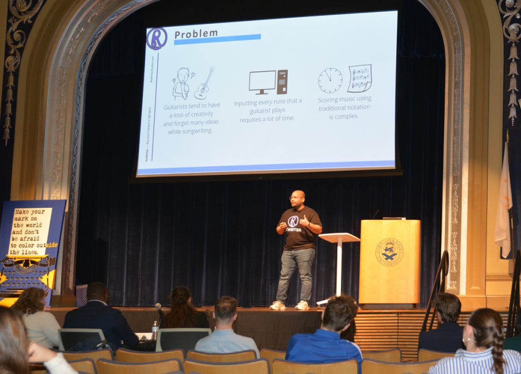 Young man, Randy Benitez is speaking on a stage in a university auditorium, while onlookers view a screen over Randy'd head displaying a slide from his presentation.