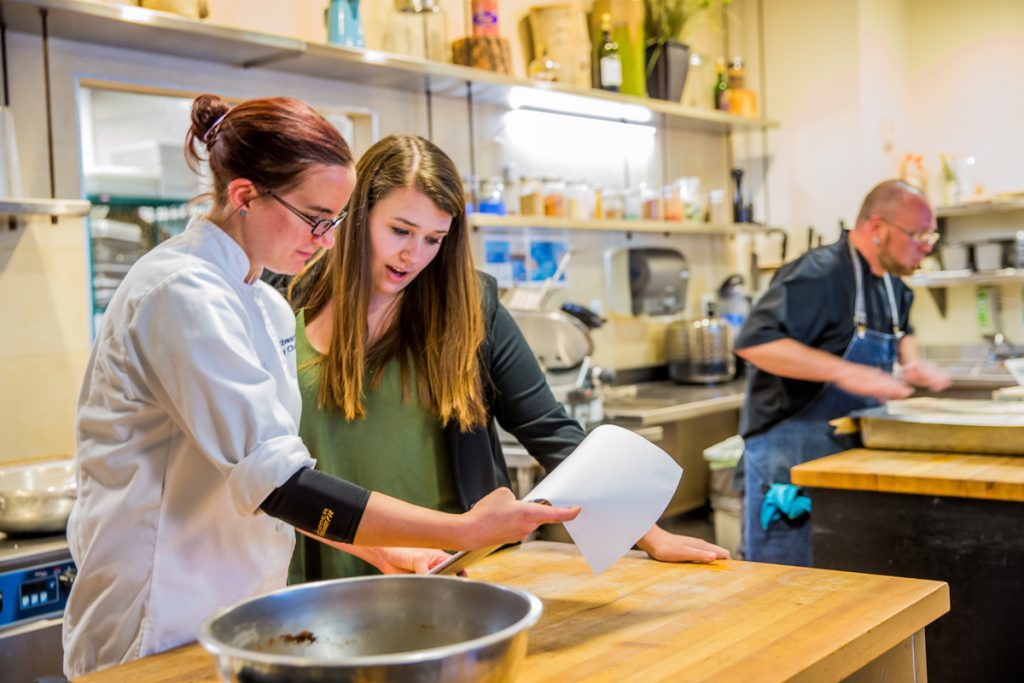 Two women in a commercial kitchen look at notes on a clipboard, while in the background a man in an apron is preparing food.