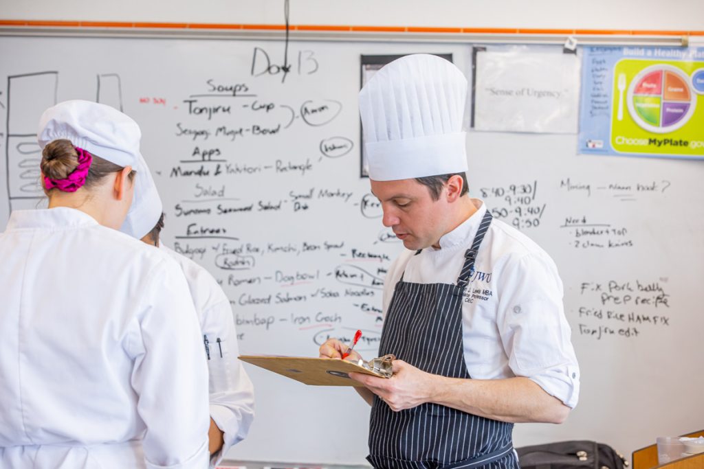 Male instructor wearing chef's cap and blue and white striped apron over white shirt embroidered with JWU insignia is holding a clipboard and making notes, while observing two students in uniforms. Behind them is a whiteboard with notes.