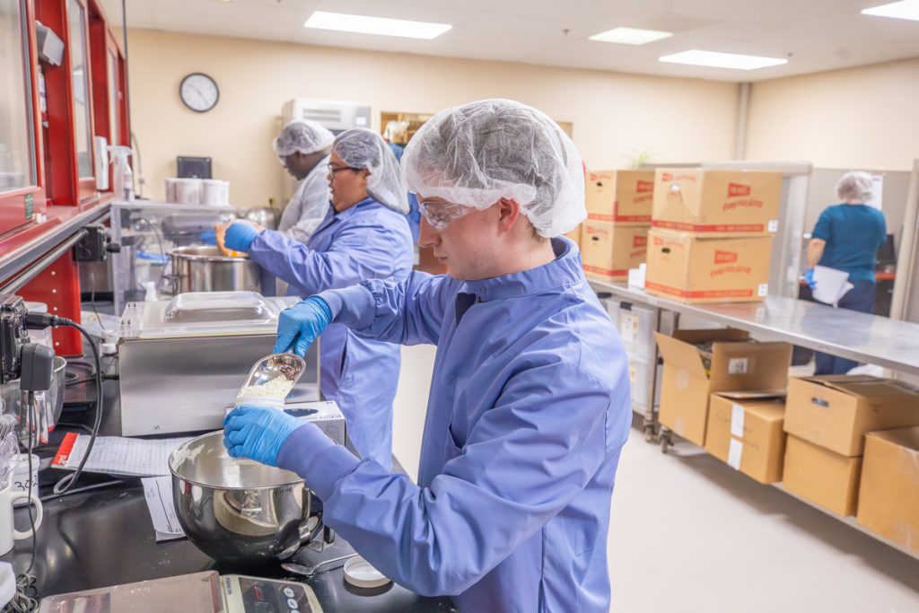 Multiple students of mixed gender, wearing blue lab coats, gloves and bouffant caps covering their hair are measuring and analyzing food products.