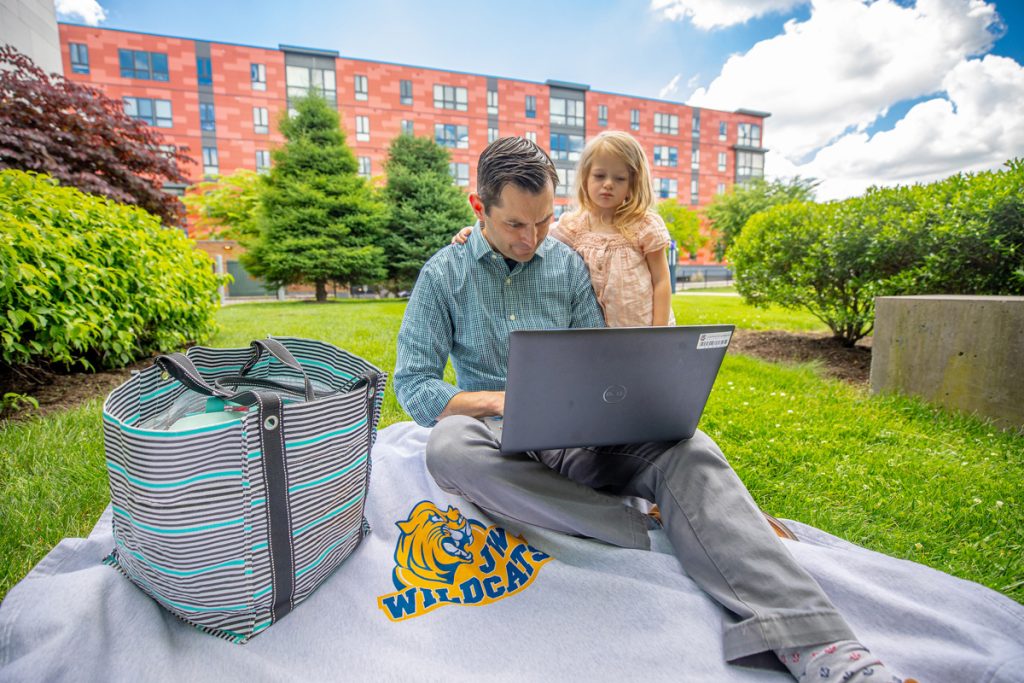 A mean is seated on the grass in an outdoor university gree space between buildings, while he and his young daughter are viewing a laptop screen together.
