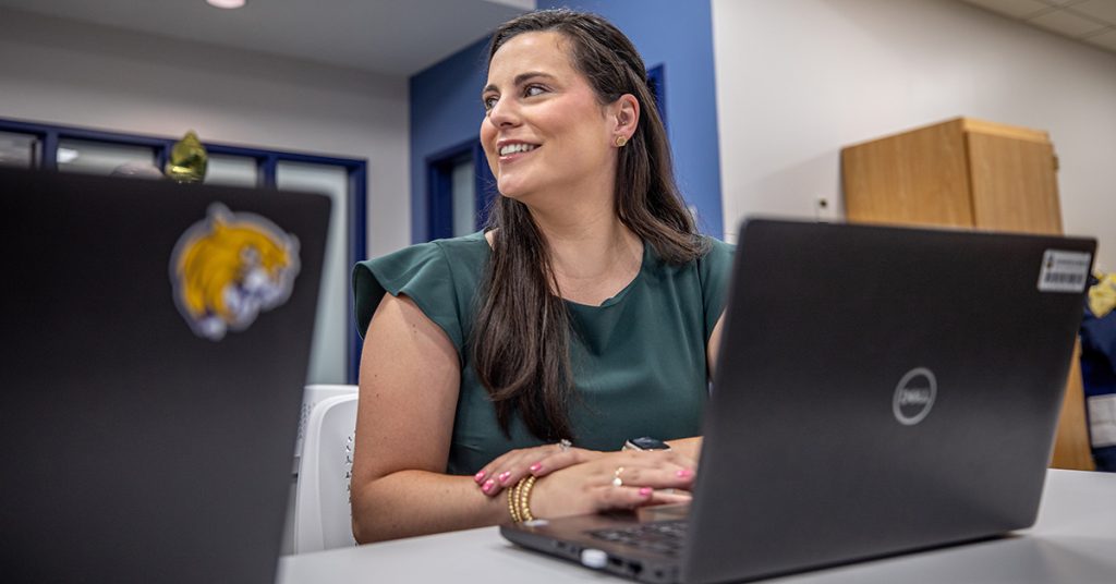 Young woman in a green dress seated at a table with her laptop looks up to greet someone in the room.
