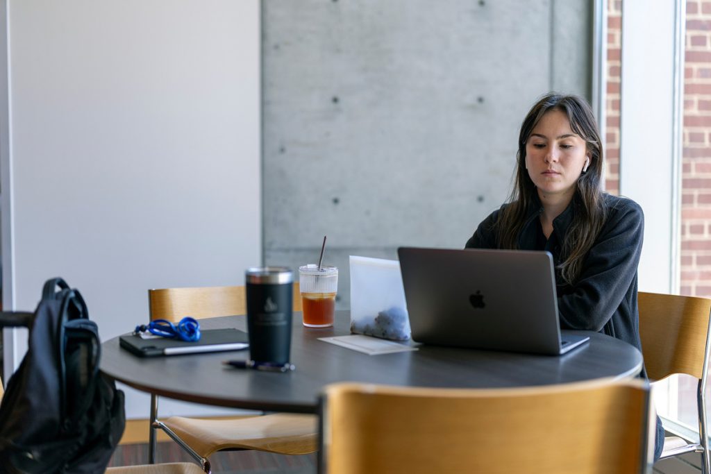 A young woman, seated at a round table on which an iced tea in a cup and a water mug are placed within reach, while she works on her laptop.