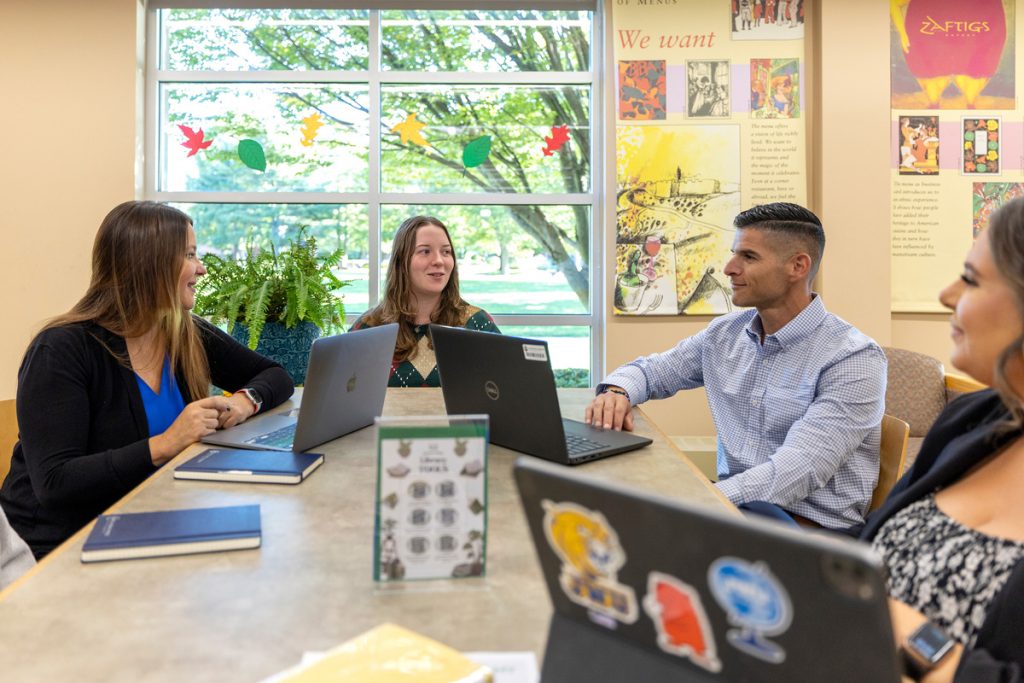 One male and three female students sitting at a table in the library, with laptops open and talking with one another.