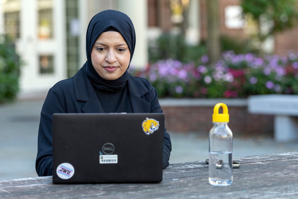 Young woman wearing a hijab, seated outdoors in a courtyard, looking at her laptop screen.