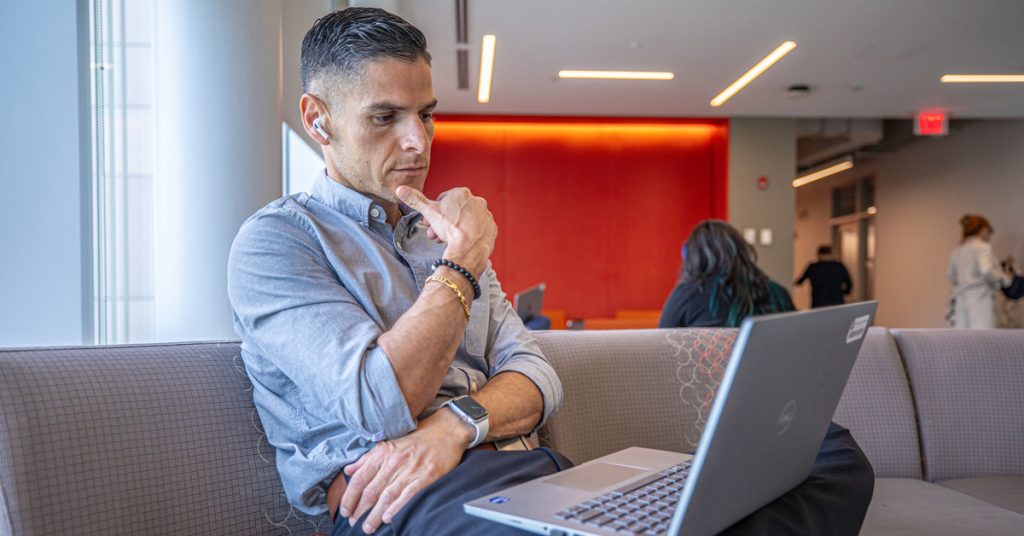 Male student seated in a common area looking thoughtful as he views his laptop screen.