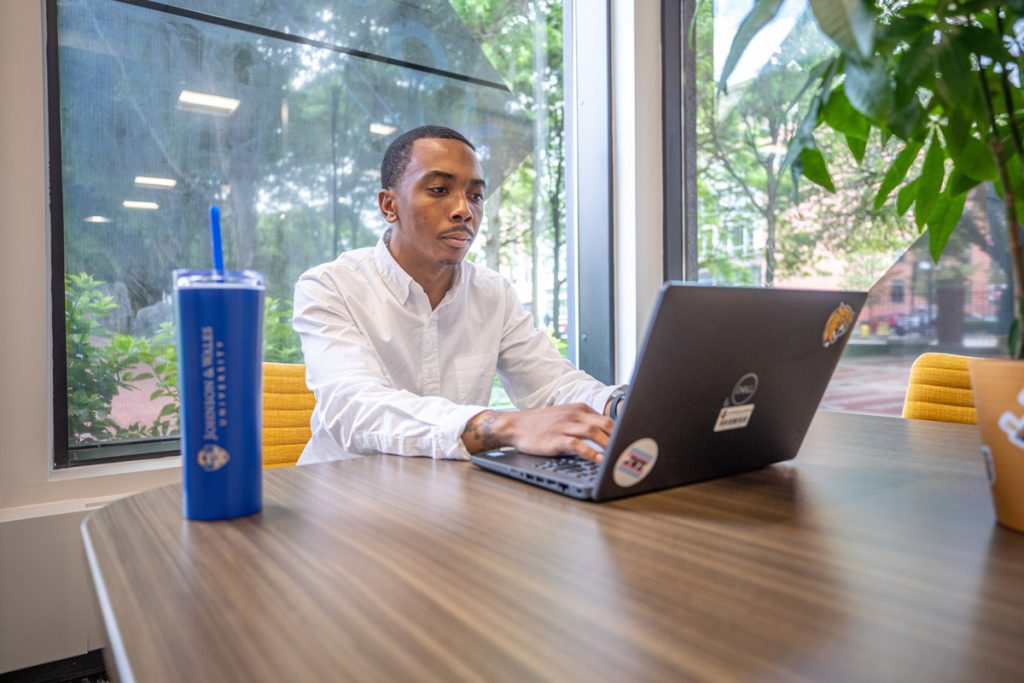 Male student works on a laptop on a table in front of large windows through which a campus courtyard can be seen.
