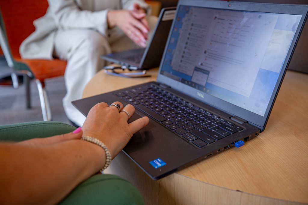 Close-up of laptop on a table displaying information on the screen and a female student's fingers on the touchpad, while in the background another student at the table is also working on a laptop.