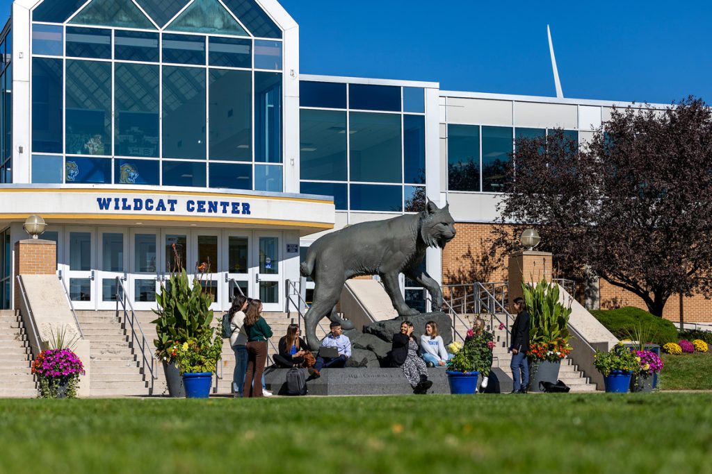 A group of students, including McKenzie Thibeault, sit on the steps in front of the Wildcat Center on the Harborside Campus.