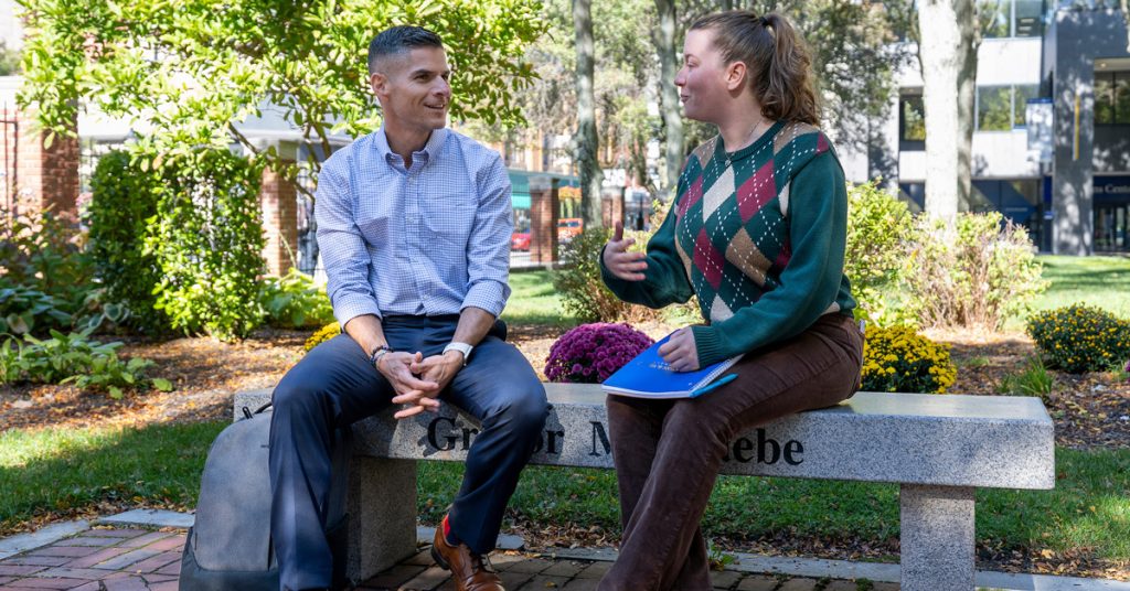 Adult male and female are seated on a stone bench in an outdoor public area, smiling and talking with one another.