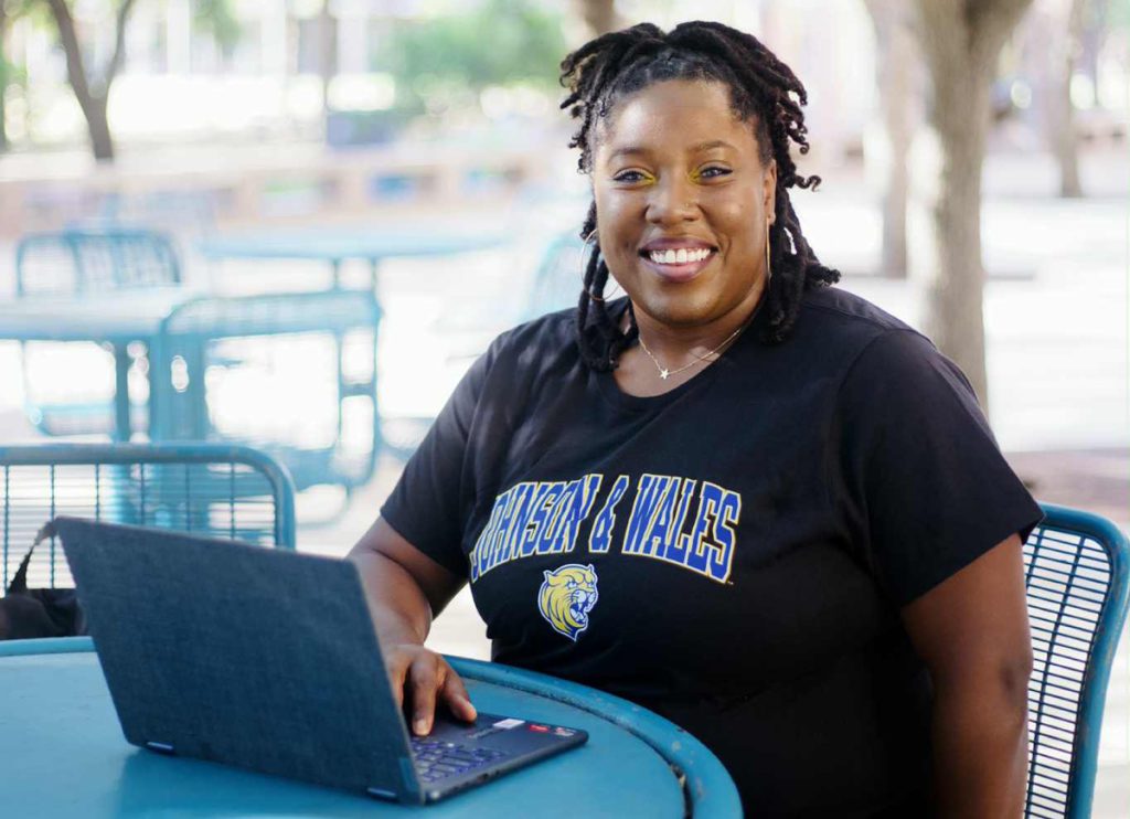 Starlisha Keith, wearing a Johnson & Wales t-shirt, is seated outdoors at a blue cafe table, while she is interning virtually on her laptop.