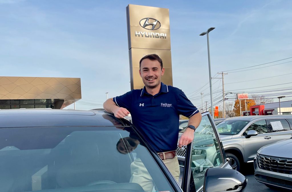 Ben Huntzinger standing in front of a Hyundai sign, surrounded by cars in the lot at Faulkner Hyundai Harrisburg where he interned and now works.