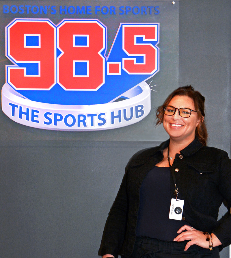 Chelsea Delfino stands alongside the 98.5 The Sports Hub logo on the wall outside the radio show's studio.