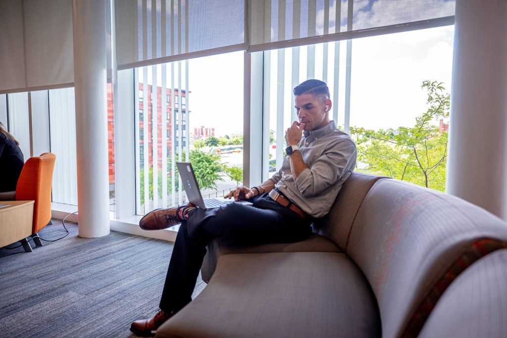 Male seated on a couch in a common area, looking thoughtful as he views a laptop screen.